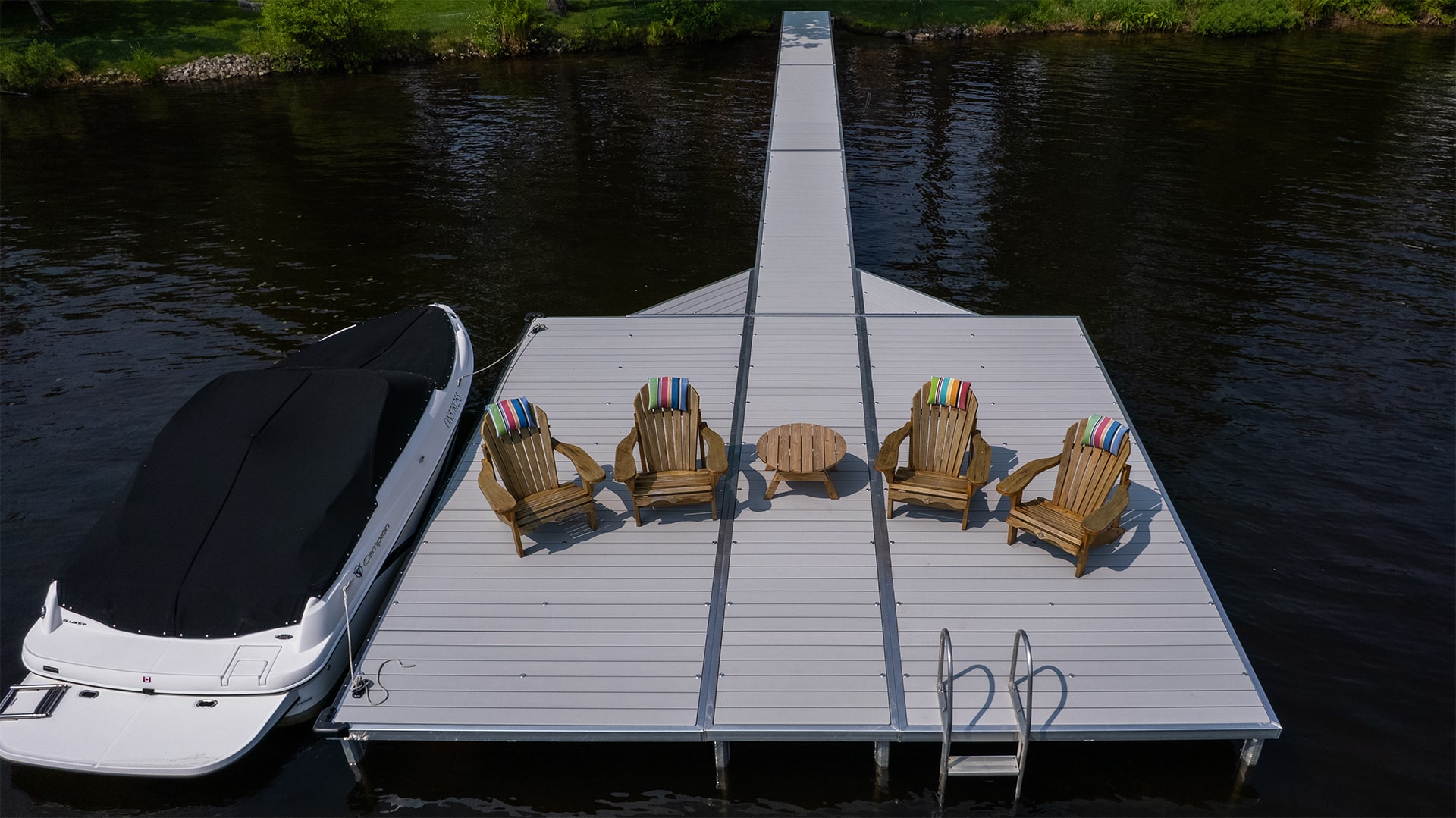 Aerial view of a lakeside dock with four wooden Adirondack chairs, a round table, and a moored boat, surrounded by calm water and greenery.